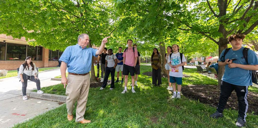 Students taking their class under green trees, the professor John Sprinkle is pointing.