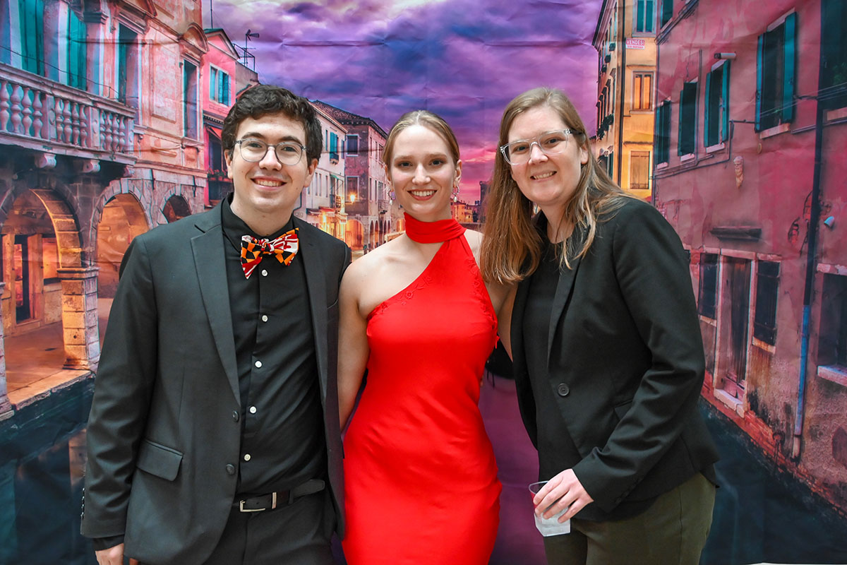 A man in a Maryland tie, a woman in a red dress and faculty member posing in front of a picture of Venice.