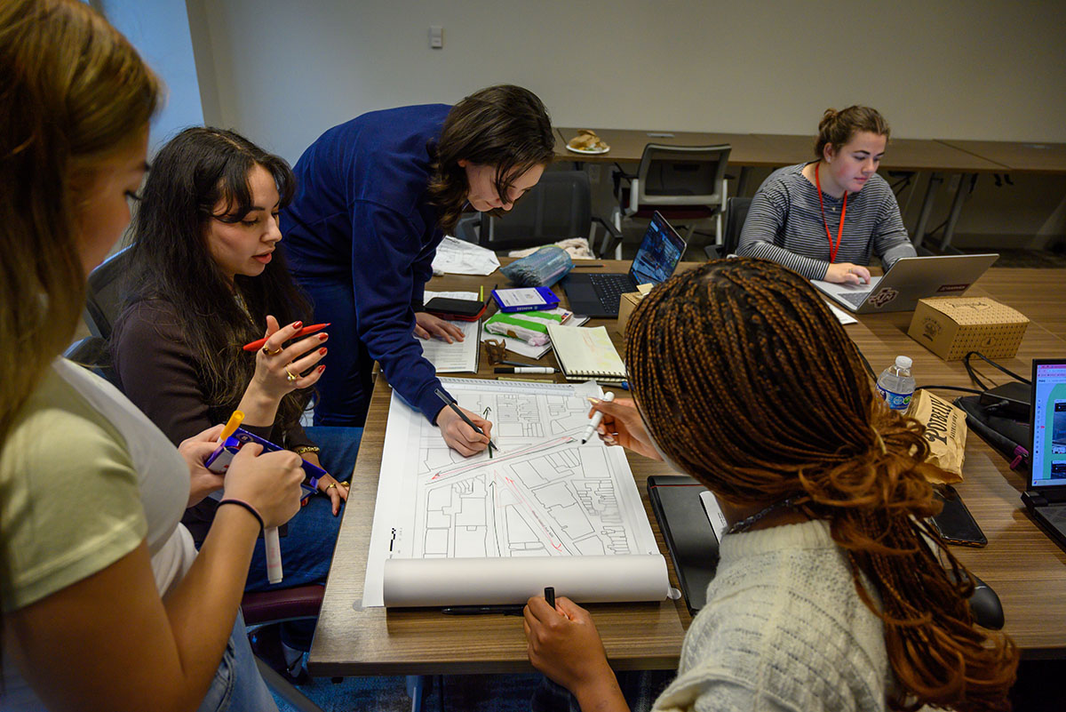 Four female students drawing on a site plan together with colorful markers. A fifth student is in the background on her laptop.