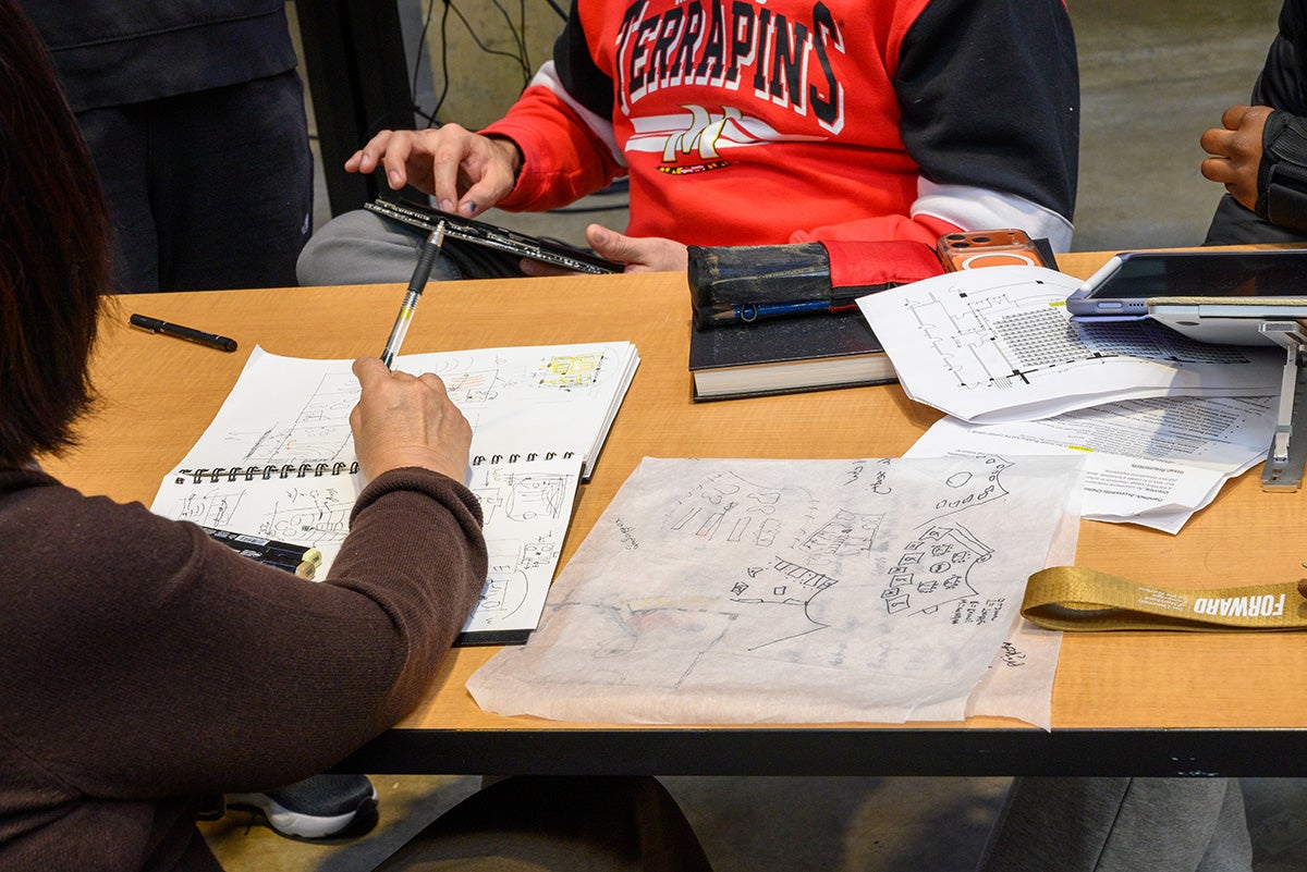 Closeup of trace paper and sketchbooks. A student with a red Terrapins shirt is zooming into a tablet in the background