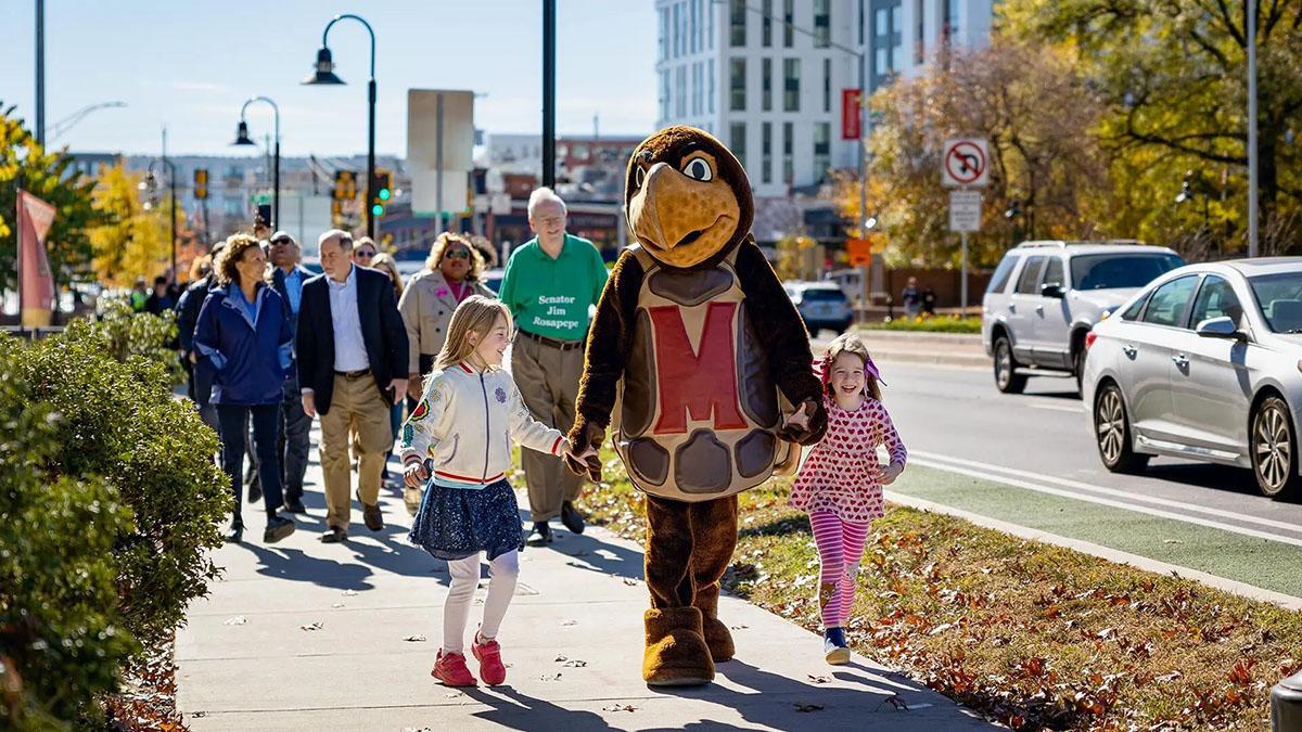 Testudo walking down main street's sidewalk with two girls and people are in the background