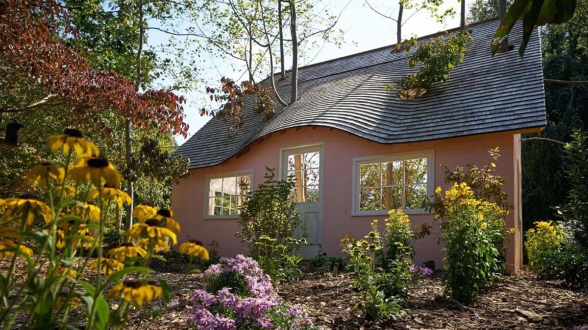 A pink house with an undulating roof, surrounded by trees and flowers