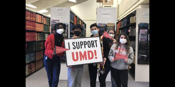 Students in the library with facemasks, holding a sign saying "I Support UMD"