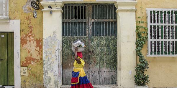 Women carrying a basket on head, walking in front of green gate in a Columbian neighborhood.