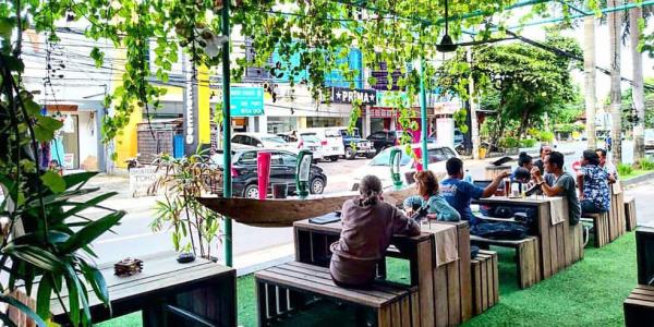 People sitting on wooden tables at a beer garden looking at cars on street