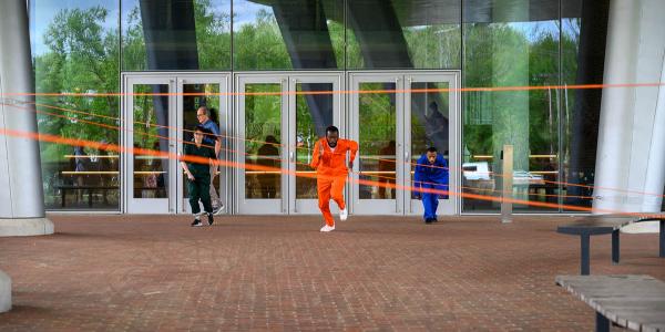 The people running toward the camera. Man in the middle is wearing an orange jumpsuit and there is orange tape serving as obstacles.