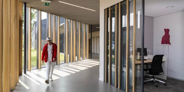 Man walking through a naturally lit hallway inside a building with tall windows and wooden structures.