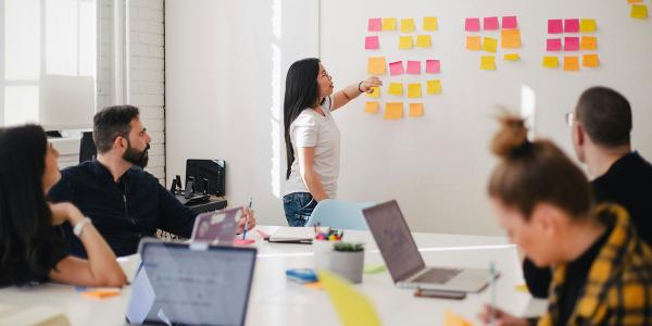 Woman pointing to sticky notes and others sitting at a table with laptops out