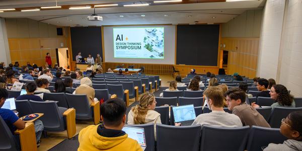 People in an auditorium talking and with their laptops open. AI+Design Symposium is on the projector