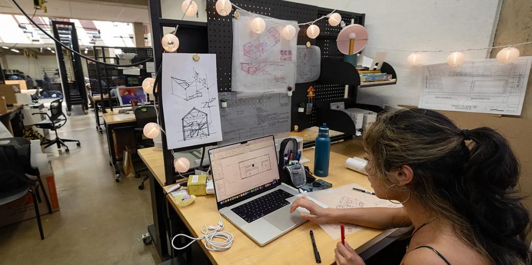 A student working on an architectural sketch on her computer, string lights and other sketches decorate her studio desk.