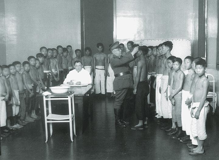 Black and white photograph of children aligned around a doctor.