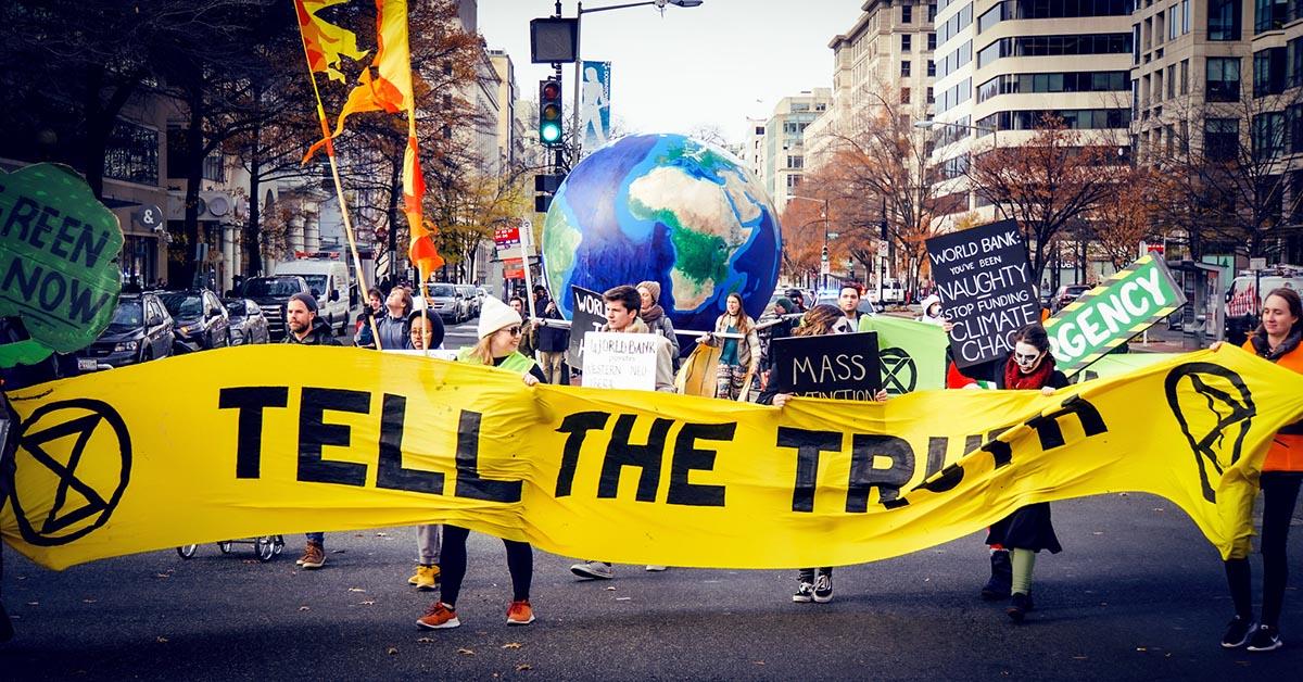 People marching and holding a yellow sign reading "Tell the Truth" a large earth globe is behind them.