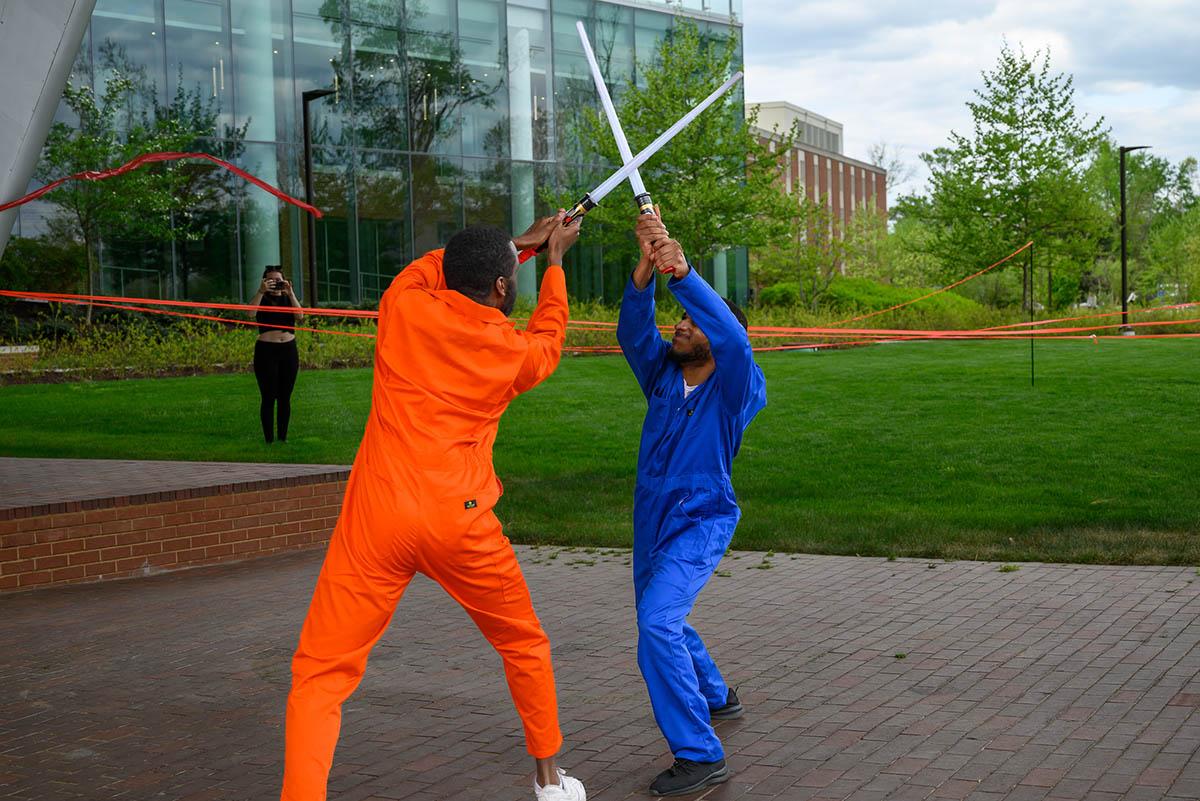 Two men play fighting with laser swords. One wears an orange jumpsuit and the other a blue one. 