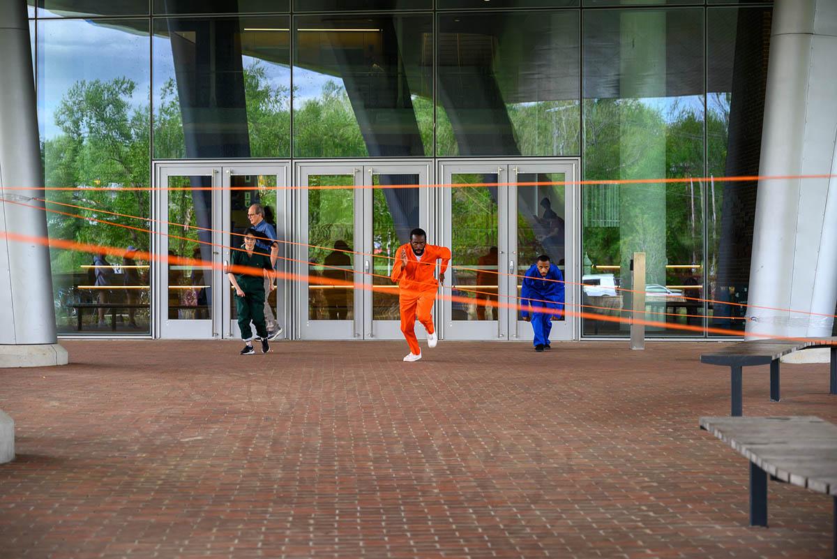 Three people running toward the camera. Man in the middle is wearing an orange jumpsuit and there is orange tape serving as obstacles.