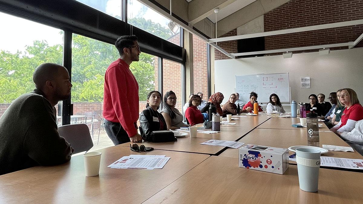 A student in red standing up at a table full of people and speaking