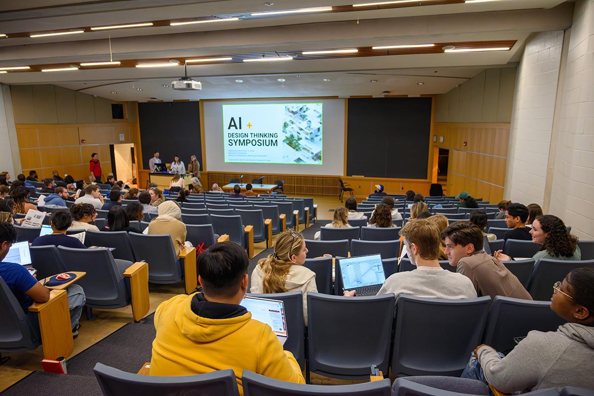 People in an auditorium talking and with their laptops open. AI+Design Symposium is on the projector