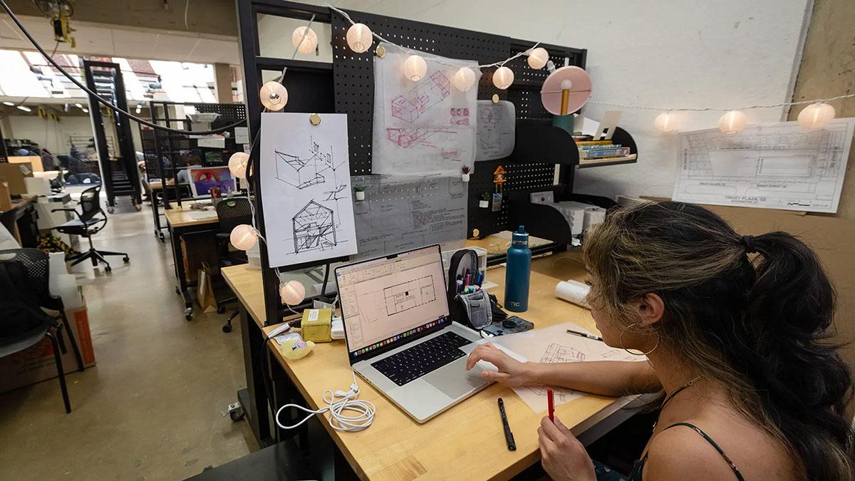 A student working on an architectural sketch on her computer, string lights and other sketches decorate her studio desk.