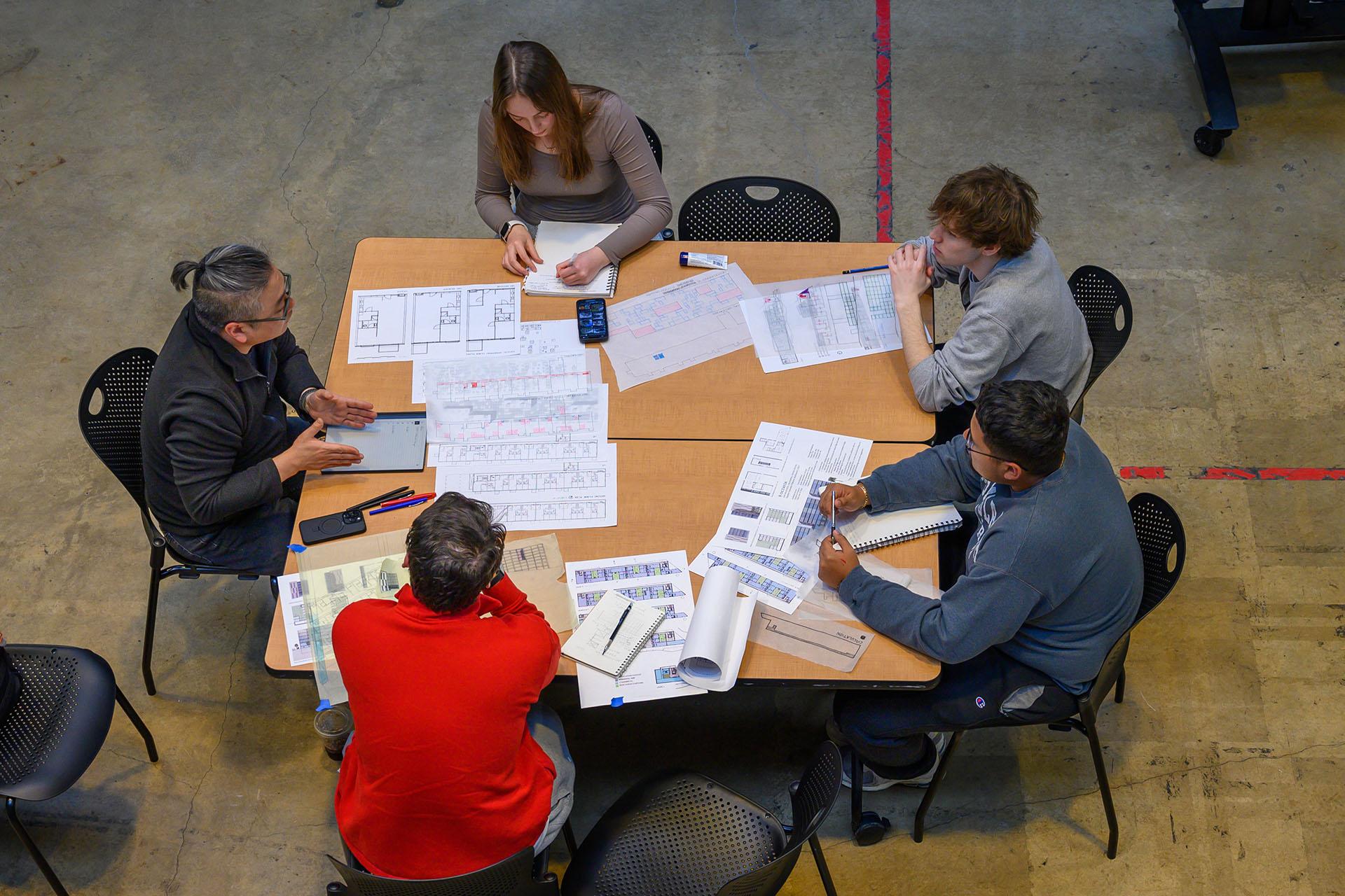 Architecture students at a table with their sketches and projects out which a professor critiques their work