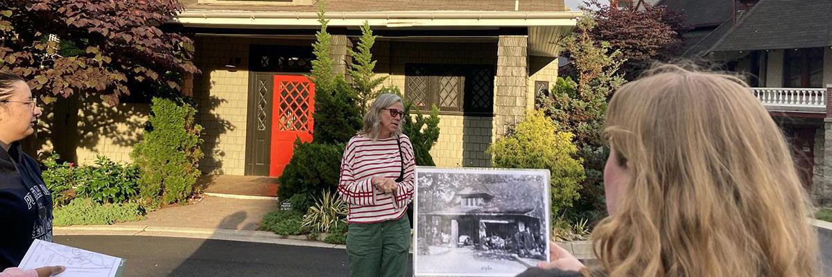 Student in foreground holding a black and white photo of a historic house, which is also in the background