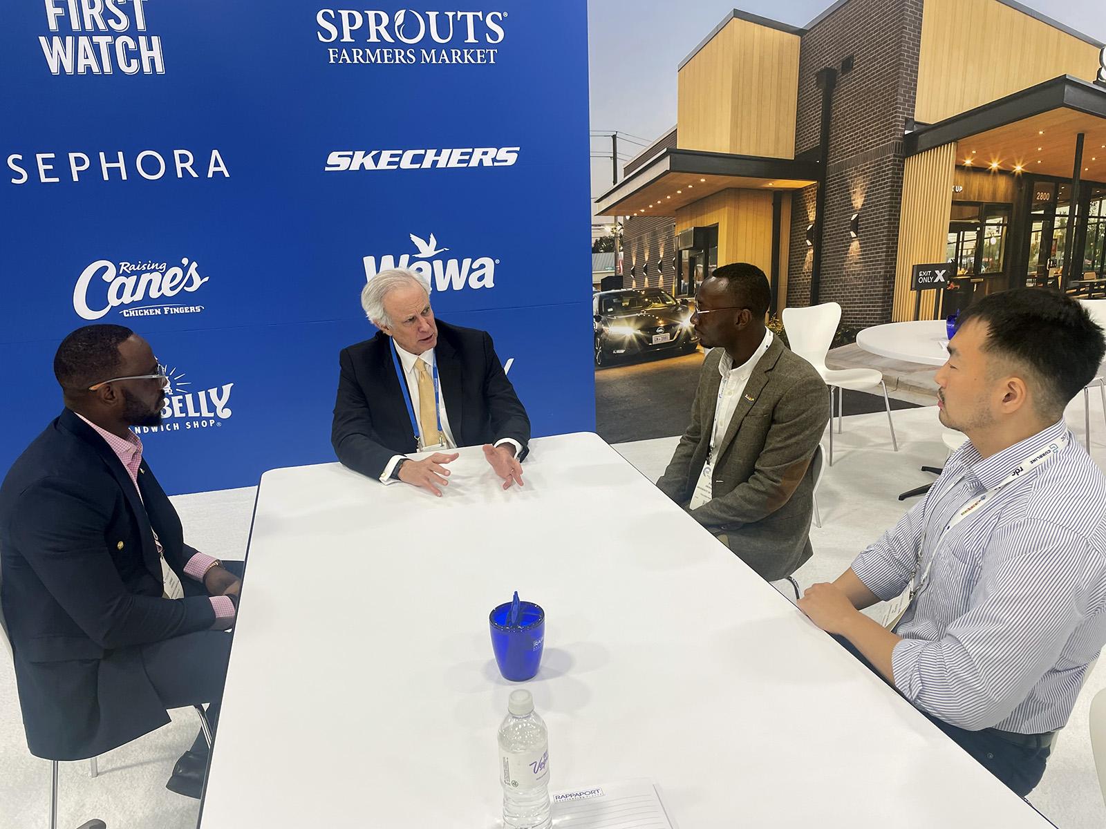 Students talking to a man on a long white table with a blue sponsor banner behind them