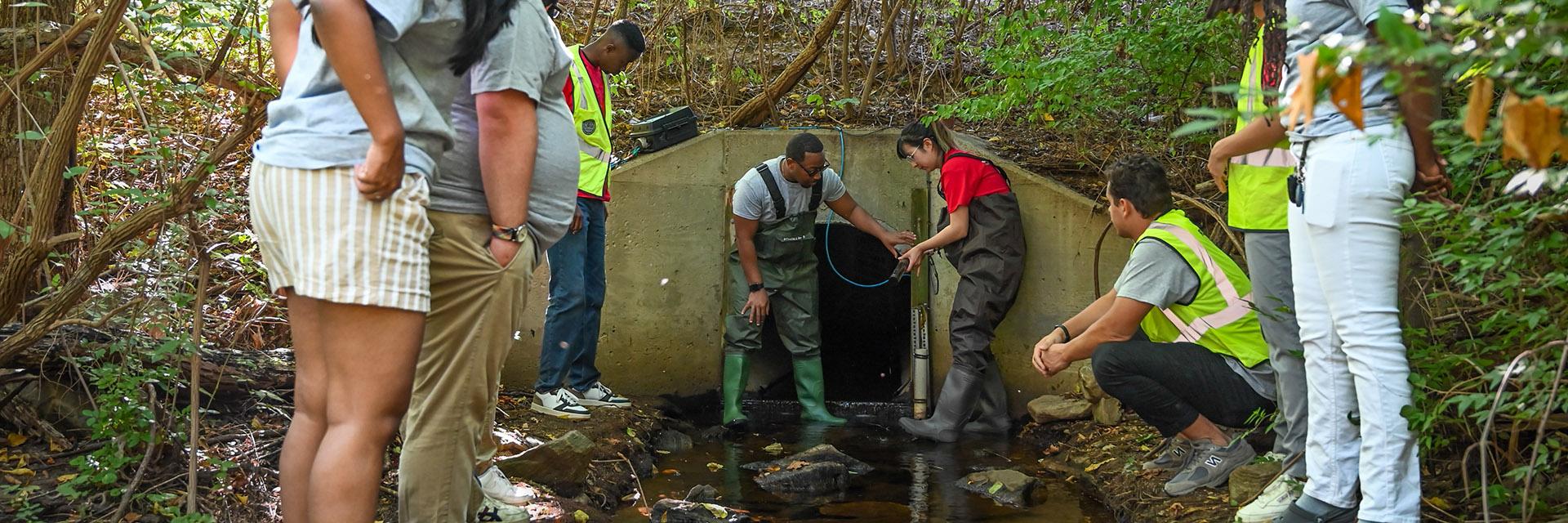 People in front a stormwater tunnel ; a man and a woman dressed in gaiters and water trousers looking at a sensor.