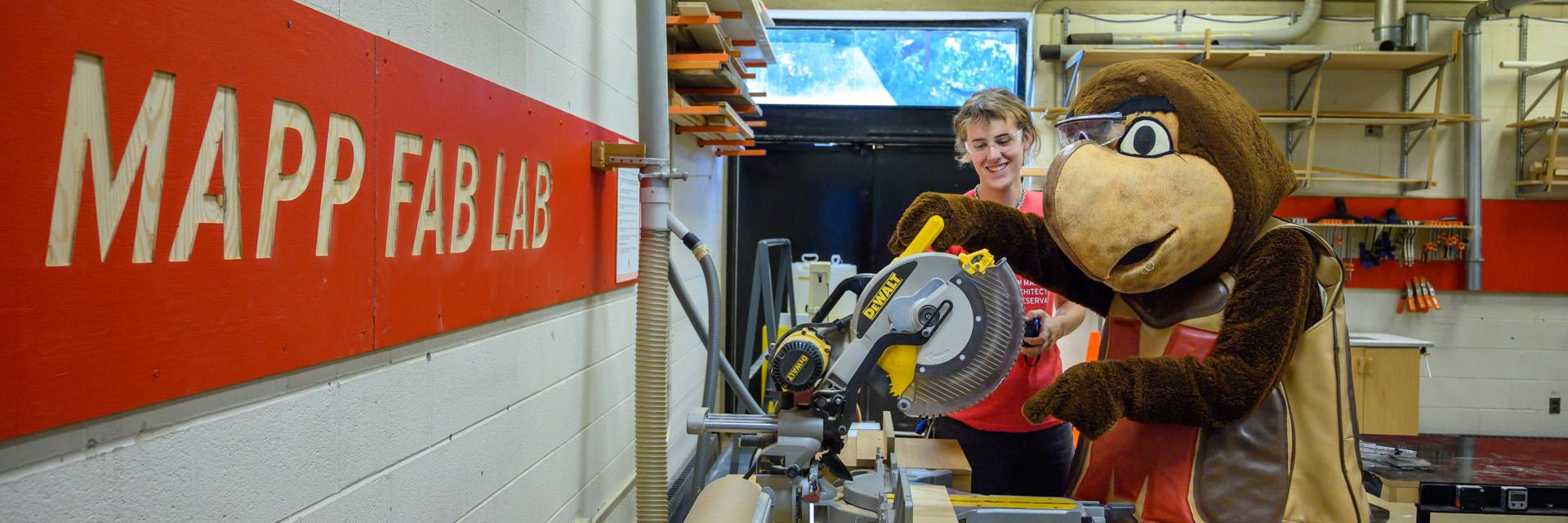 Testudo testing a hand saw at the MAPP Fab Lab, with shop technician. Both are wearing safety glasses.