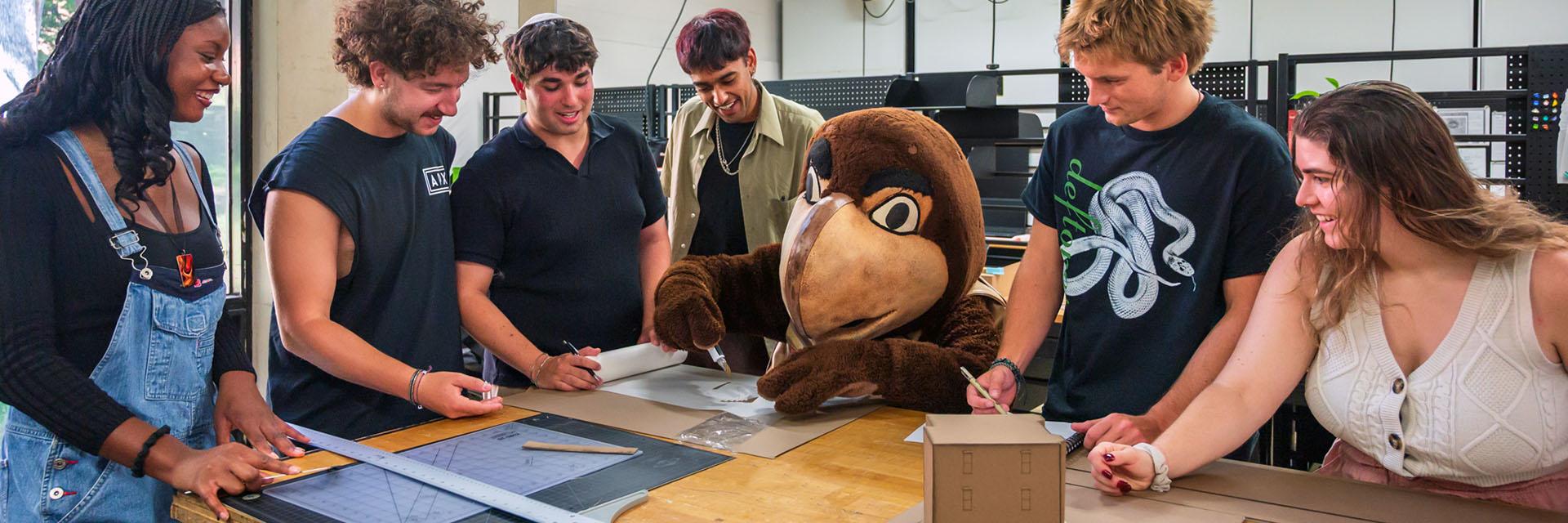 Testudo working on cutting paper surrounded by students in the architecture studio