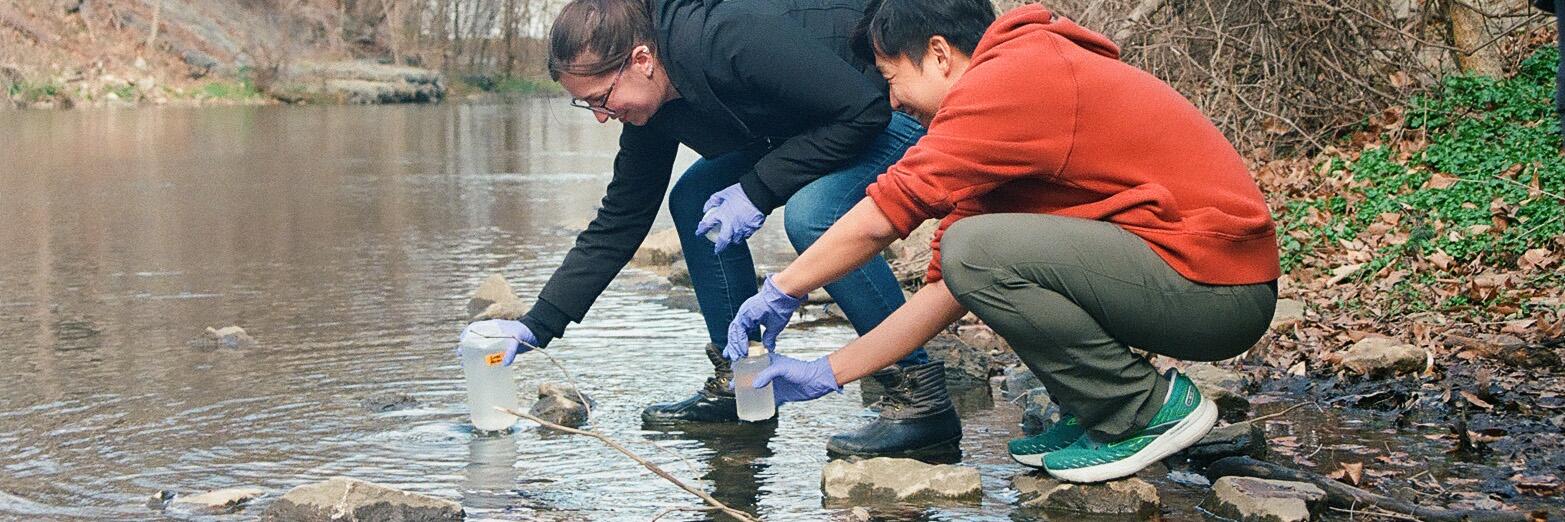 A man and a woman standing by water and testing it. They are wearing plastic gloves and collecting samples