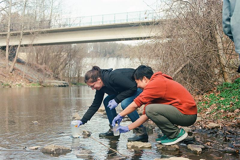 A man and a woman standing by water and testing it. They are wearing plastic gloves and collecting samples