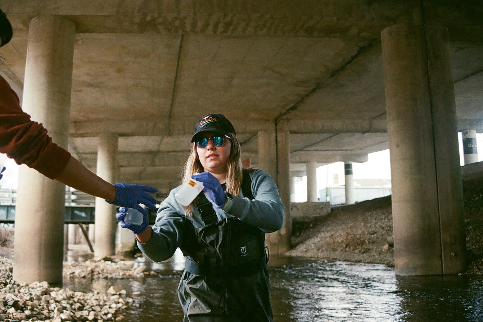 A hand is handing water sampling bottles to a woman wearing sunglasses glasses, who is under a bridge