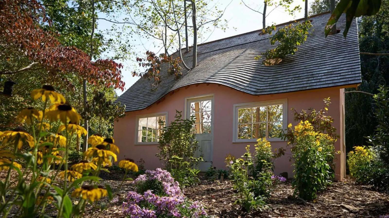 A pink house with an undulating roof, surrounded by trees and flowers