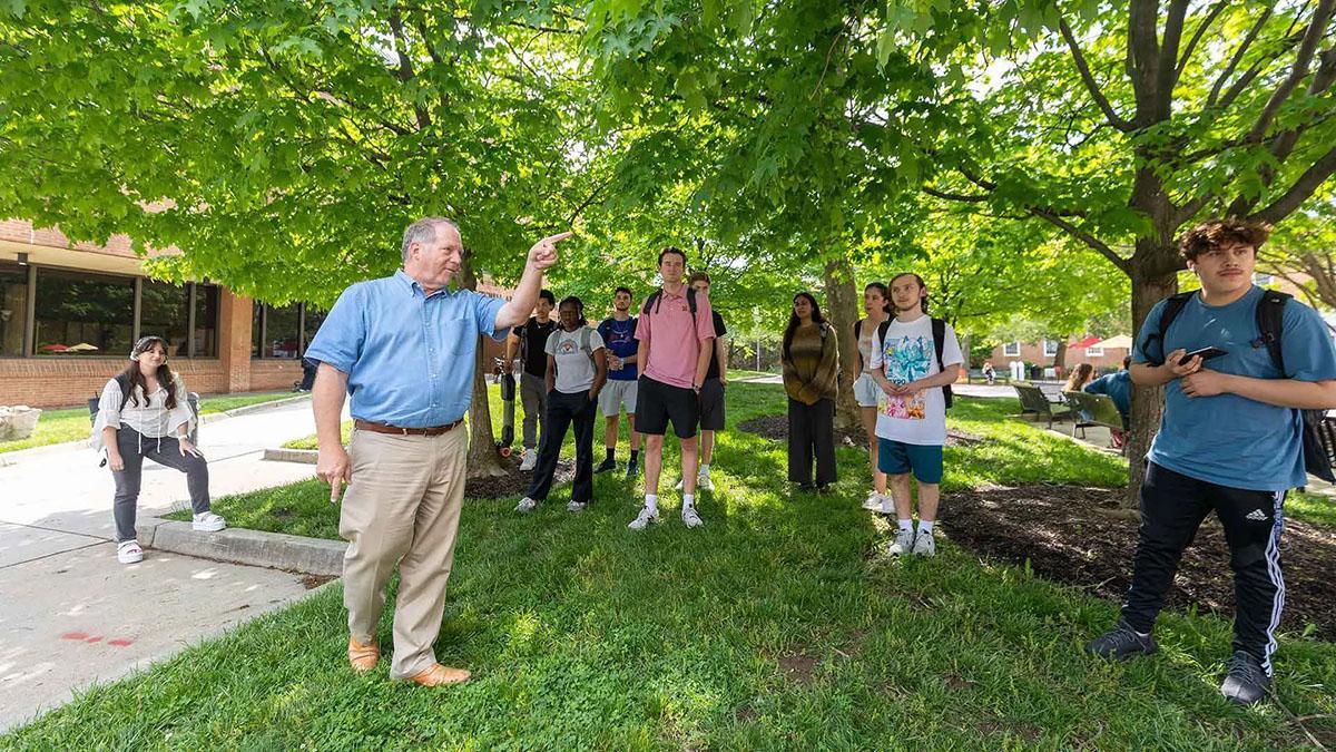 Faculty pointing upward with students around him, they are surrounded in greenery and trees.