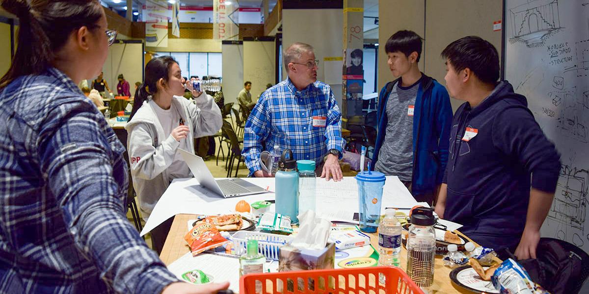 Students and mentor gathered around a table with computers and plans