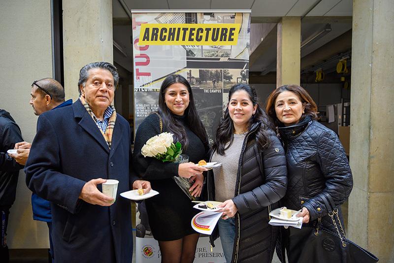 A graduating student holding flowers and posing with her family by an Architecture banner