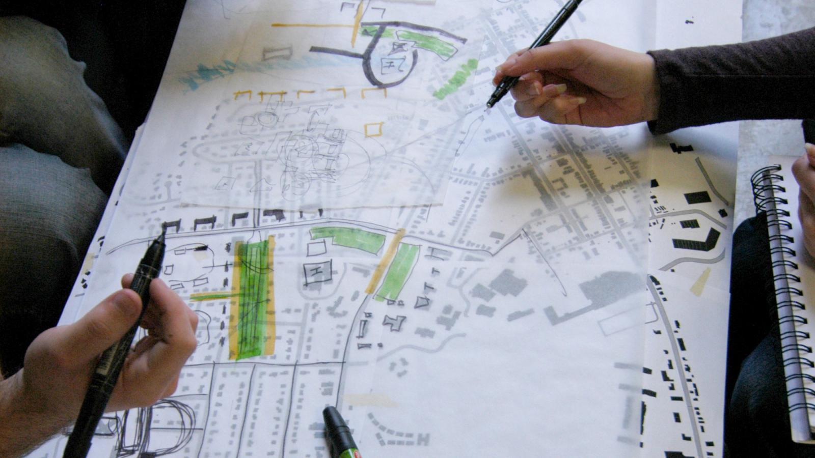 Two people’s hands hold pens and point to a large printed map covered with sketches, notes, and colored highlights, suggesting they are planning or discussing a design or development layout.