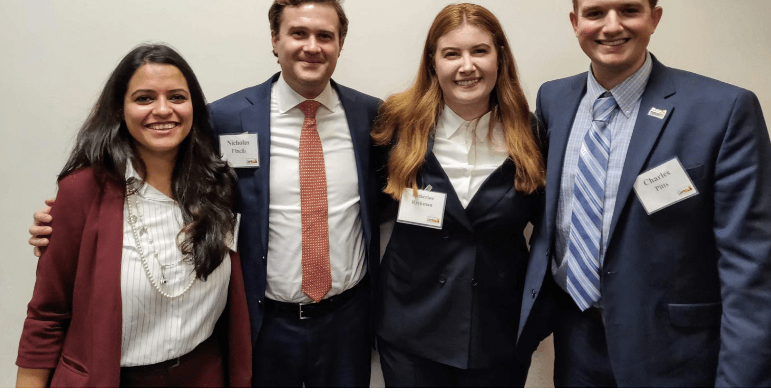 Four young adults in business attire stand close together, smiling at the camera in front of a plain wall.