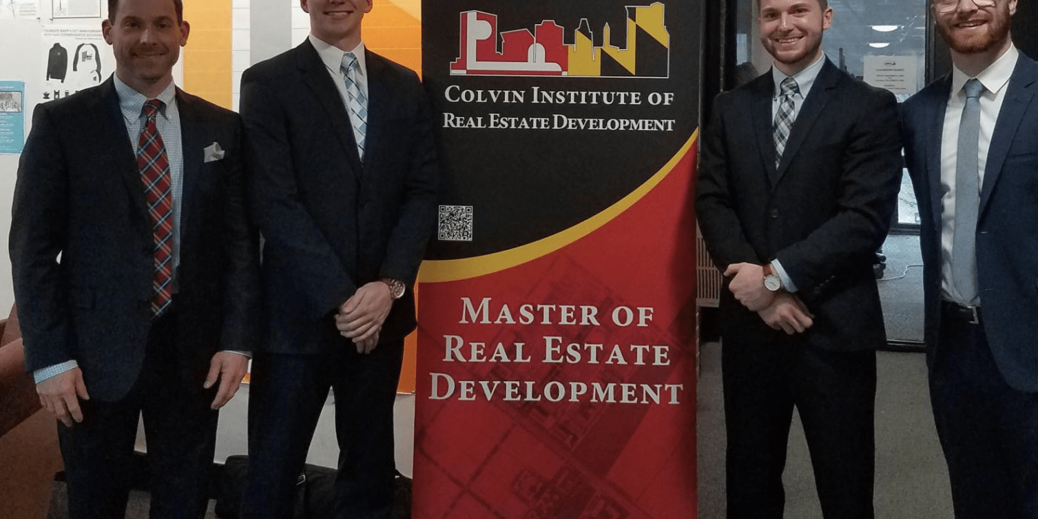 A group of four men in business suits stand indoors on either side of a tall banner that reads “Colvin Institute of Real Estate Development” and “Master of Real Estate Development.” 