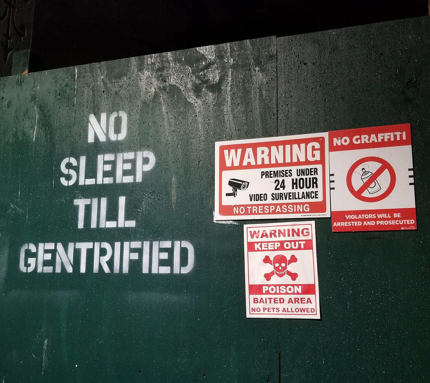 Graffiti writing saying "No Sleep Till Gentrified" by signage that states no graffiti and that the location is under surveillance