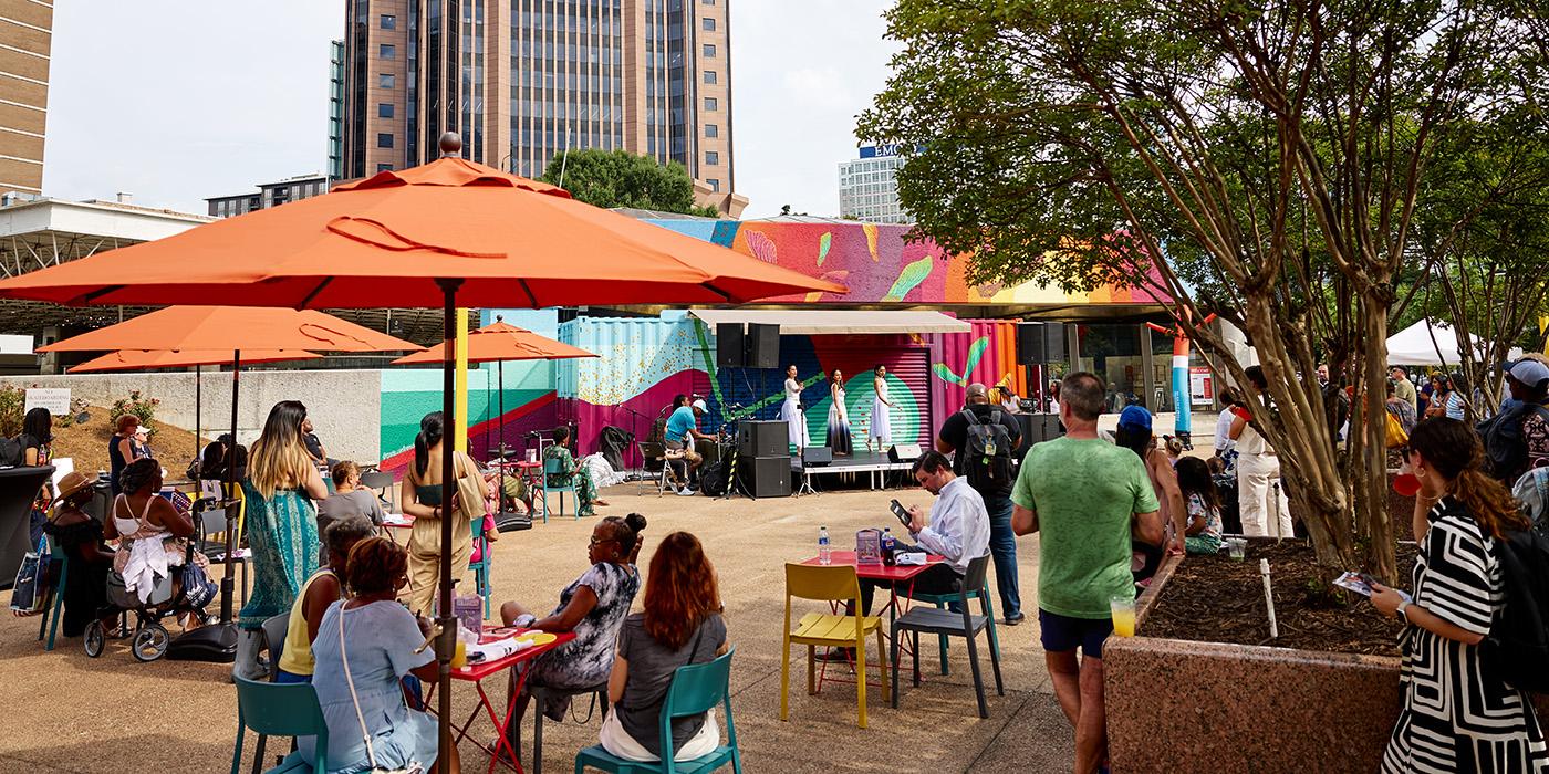 People sitting on colorful chairs under umbrellas at a plaza. Three women are dancing on stage in front of a colorful container. 
