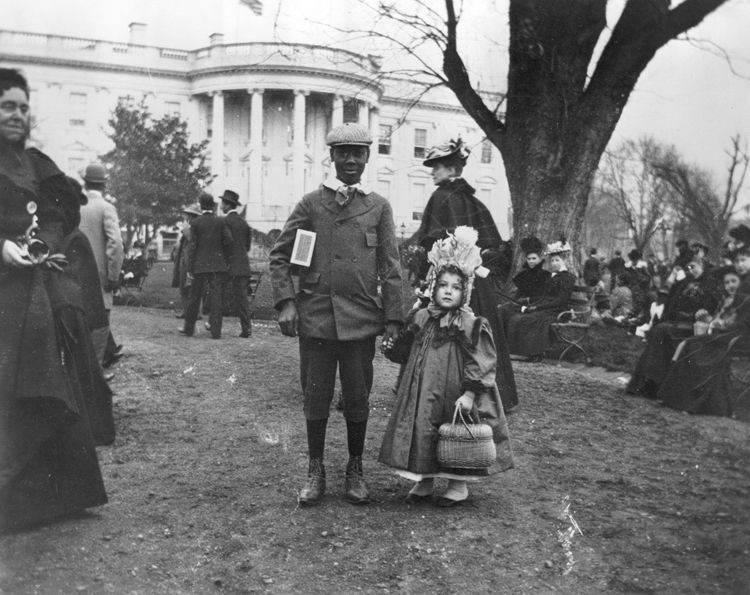 A little girl dressed for the White House egg roll, holding a boys hand, in front of the White House